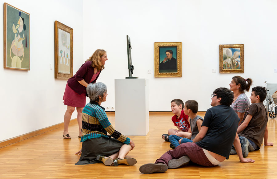 Un groupe d'enfants en situation de handicap, assis dans les salles d'art moderne avec deux médiatrices du musée